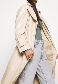Beige Damen-Trenchcoat offen getragen, grünes geripptes Langarmoberteil und hellgraue hochgeschnittene Jeans vor neutralem Hintergrund.