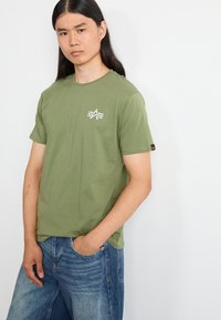 Olivengrøn bomuld T-shirt med korte ærmer og en rund halsudskæring. Inkluderer et lille hvidt grafisk motiv på brystet og afslappede denimjeans.