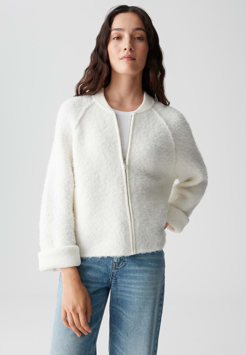 Witte, fluffy zip-up jas met een geribde kraag en boorden. Heeft raglanmouwen en een zachte textuur, gecombineerd met een blauwe spijkerbroek.