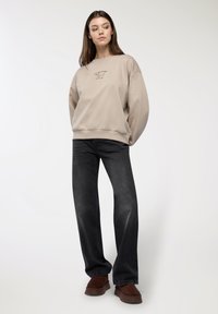 Beżowy sweter z nadrukiem graficznym, luźny krój i ściągacz na dole. Połączony z ciemnoszarymi spodniami dzwonami i brązowymi zamszowymi mokasynami.