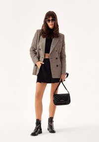 Blazer de cuadro de doble botonadura en tonos marrones, top corto negro, falda corta, botas negras brillantes hasta el tobillo con detalles metálicos y un bolso negro acolchado.