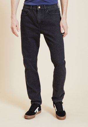 Jeans en denim noir avec une coupe droite, dotés de cinq poches, d'un bouton en métal et de coutures discrètes. Portés avec des baskets en daim noir.