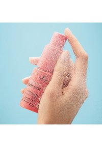 Rosa zylindrische Sprühflasche mit der Aufschrift "Soft Dust", die in einer Hand gehalten wird und feinen Staub versprüht, vor einem hellblauen Hintergrund.