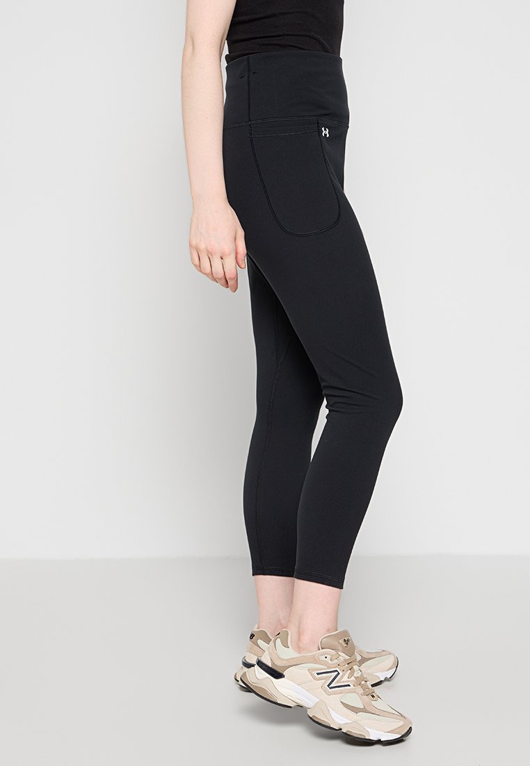 Leggings neri a vita alta, realizzati in tessuto elasticizzato. Presentano una tasca laterale e una texture liscia, abbinati a scarpe da ginnastica beige chiaro.