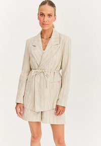 Pruhovaný blazer a odpovídající šortky v světlé béžové barvě s pastelovými akcenty. Měkká tkanina, přední kapsy a detaily se zavázáním v pase.