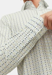 Chemise à manches longues en tissu léger, avec une base blanche ornée de motifs de feuilles verts, bleus et marine. Comprend des poignets à boutons et un col.