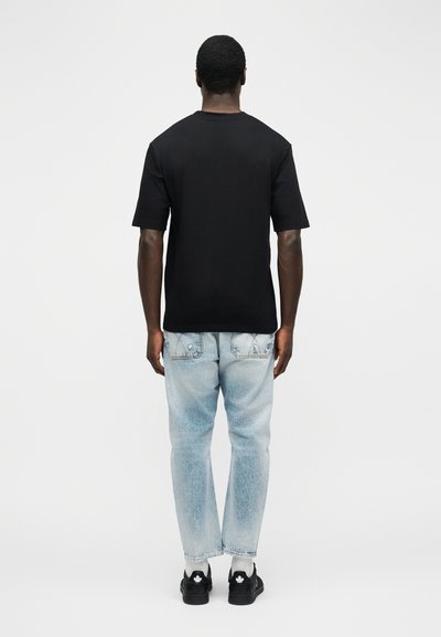 Άνδρας που κοιτάζει προς τα πίσω, φοράει απλό μαύρο μπλουζάκι, ανοιχτόχρωμο τζιν, λευκές κάλτσες και μαύρα αθλητικά παπούτσια με λευκά λογότυπα φύλλου.