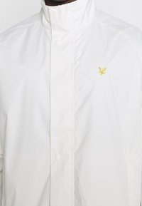 Biała kurtka z wysokim kołnierzem i zapięciem na zatrzaski, z małym żółtym haftowanym logo ptaka na piersi.