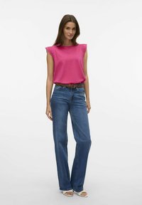 Roze mouwloze top met kantaccenten, gecombineerd met lichtblauwe wijde jeans en witte sandalen. Model staat voor een effen achtergrond.