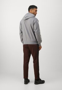 Szürke kapucnis pulóver, bordás mandzsettákkal, barna, oldalcsíkos fekete csíkkal ellátott szűkített nadrággal párosítva. A megjelenést fekete sportcipő teszi teljessé.