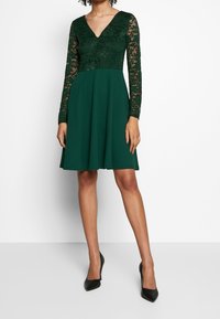 Grünes Kleid mit einem Spitzenoberteil, langen Ärmeln und einem V-Ausschnitt, ausgestelltem Rock aus strukturiertem Stoff, kombiniert mit schwarzen Schuhen.