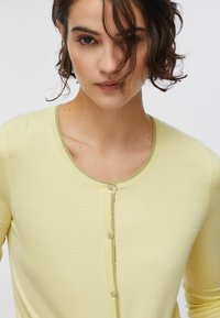 Rövid, göndör sötét hajú nő, aki halvány sárga kardigánt visel virág alakú díszgombokkal, fehér háttér előtt.