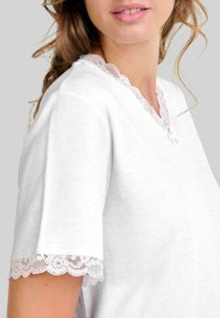 Mujer vistiendo una blusa blanca de mangas cortas con encaje en las mangas y el escote, de tela suave, piel clara, cabello suelto y ondulado sobre un fondo liso.