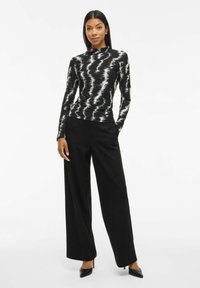 Femme portant un haut à manches longues noir et blanc à motifs, un pantalon noir à larges jambes et des escarpins noirs à bout pointu, se tenant les mains dans les poches.
