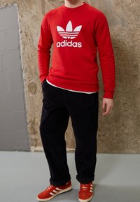 Röd sweatshirt med vitt Adidas-logotyp, matchad med mörkblå byxor och röda skor med vita ränder. Slät tyg, avslappnad passform.