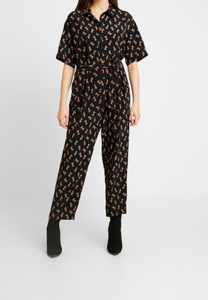 Kvinde iført en sort jumpsuit med korte ærmer og et lille orange dyreprint, kombineret med sorte ankelstøvler.