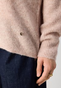 Helles rosa, flauschiges Sweatshirt mit gerippter Textur, versehen mit einem kleinen goldenen Logo-Button. Kombiniert mit dunkelblauen Jeans.