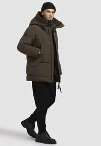 Polstret olivengrøn parka med høj krave, lynlåslommer og sorte detaljer. Kombineret med sorte bukser og robuste sorte støvler.