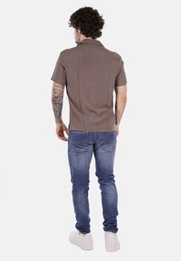 Camicia a maniche corte con colletto in tessuto marrone chiaro, abbinata a jeans blu. La camicia presenta una texture liscia e dettagli di cucitura minimi.