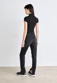Svart ribbet topp med høy hals, parret med høyt liv mørk denim jeans og svarte joggesko. Modellen står med ryggen til mot en nøytral bakgrunn.