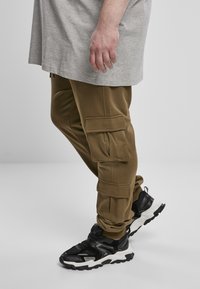 Oliven grønne cargo-bukser med flere lommer, parret med sorte sneakers med hvide accenter og teksturerede såler. Grå t-shirt ovenfor.