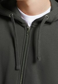 Sudadera con cremallera de color verde oscuro y textura de tela lisa, con herrajes de cremallera plateados y cordones, sobre una camiseta blanca.