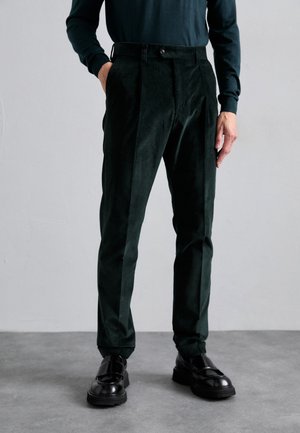Homme portant un pantalon en velours côtelé noir, des mocassins noirs et une chemise à manches longues foncée, se tenant avec une main dans la poche sur un sol gris.