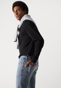 Zwarte longsleeve gemaakt van zachte stof, met een lichtgrijze sweatshirt over de schouders; gecombineerd met lichtblauwe jeans.