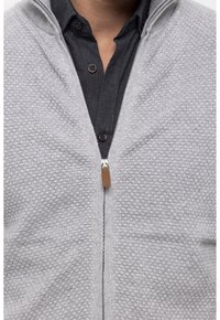 Grau strukturierter Reißverschluss-Pullover mit hohem Kragen und Leder-Ziehschlaufe, kombiniert mit einem schwarzen Hemd mit Knöpfen.