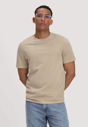 Junger Mann, der klare Brillen, ein beiges kurzärmliges T-Shirt und blaue Jeans trägt, steht mit den Händen hinter dem Rücken vor einem schlichten Hintergrund.