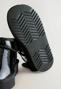 Czarne lakierowane buty skórzane z teksturowaną czarną gumową podeszwą z zygzakowatym wzorem, z widocznym szczegółowym przeszyciem wzdłuż górnej krawędzi.