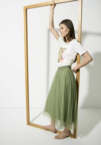 White T-shirt with a colorful graphic, green tulle maxi skirt, and beige flats adorned with studs. Model stands framed in wood.