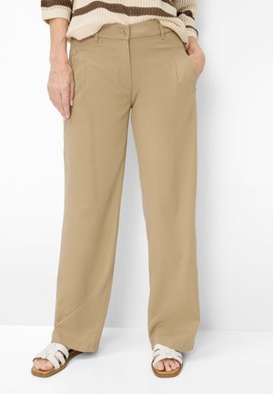 Kvinde iført beige bukser med vide ben, en stribet strikket sweater og hvide sandaler med åben tå, stående mod en hvid baggrund.