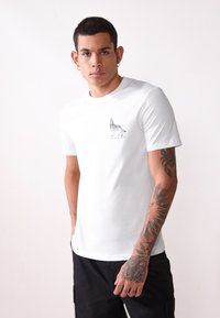 Camiseta de algodón blanca con un gráfico de un lobo negro y el texto "WALK ALONE" en el lado izquierdo del pecho. Mangas cortas, cuello redondo, textura suave.