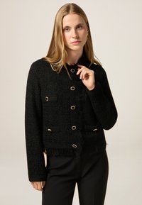 Giacca nera texturizzata con orlo a frange, bottoni color oro e due tasche anteriori. Realizzata in tessuto tessuto con una silhouette aderente.