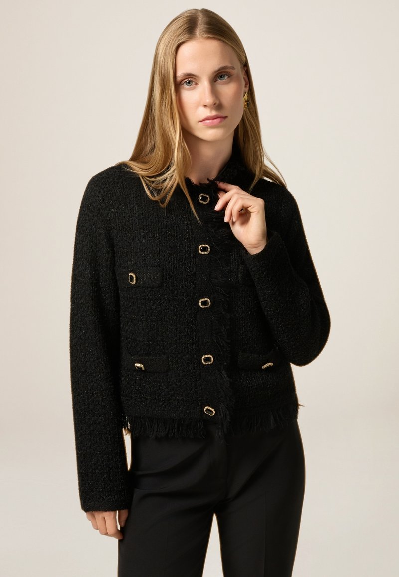 Giacca nera texturizzata con orlo a frange, bottoni color oro e due tasche anteriori. Realizzata in tessuto tessuto con una silhouette aderente.