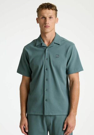 Young man wearing teal short-sleeve button-up shirt with a small triangle logo on the left chest, standing against a plain white background.