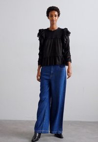 Black ruffled blouse with long sleeves paired with wide-leg blue denim pants. Model stands on a neutral background, wearing black boots.