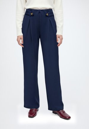 Personne portant un pantalon large bleu marine avec des détails de boutons à la taille et des mocassins en cuir bordeaux.