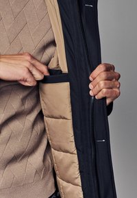 Puffatakki, jossa on tummansininen ulkopinta ja beige sisävuori, vasemmalla puolella tasku ja päällä teksturoitu neulepaita.