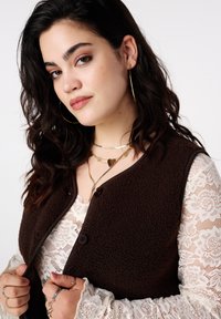 Bruin fleecevest met knoopsluiting over een wit kanten topje. De outfit wordt gecombineerd met gelaagde gouden kettingen en zilveren ringen. Krullend, donker haar.