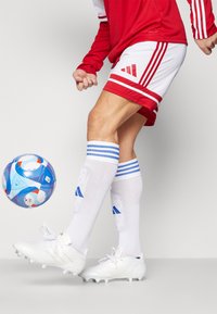 Adidas futballmez vörös mezből, fehér rövidnadrágból, fehér zoknikból kék csíkokkal és fehér cipőkből áll. Egy színes focilabda van a közelben.