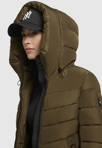 Doudoune vert olive avec une grande capuche, fermeture éclair et marquage noir. Elle présente une texture lisse et une casquette noire amovible.