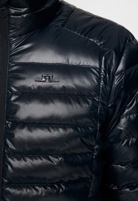 Lähikuva mustasta vatitud takista, jossa on horisontaalinen ommel ja väike metalline logo vasakul rinnal musta tõmbluku lähedal.