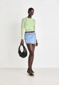 Lys grønn langermet topp med teksturert mønster, parret med en lys blå mini denimskjørt. Modellen holder en svart håndveske. Svart kilehæler.