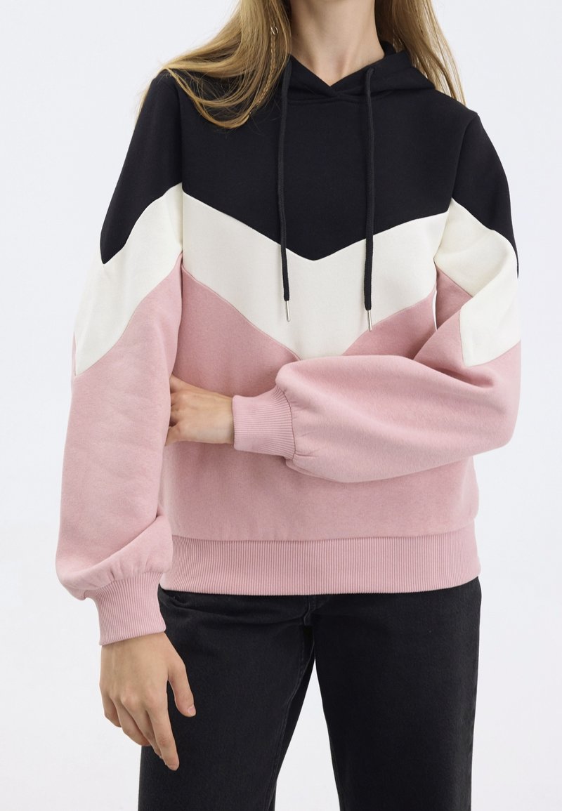 Femme portant un sweat à capuche color-block avec des sections noires, blanches et roses, associée à un pantalon noir, debout devant un fond uni.