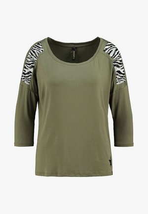 Oliven grøn skjorte med trekvartærmer, med sorte og hvide zebra-mønstrede palietter på skuldrene. Blødt stof.