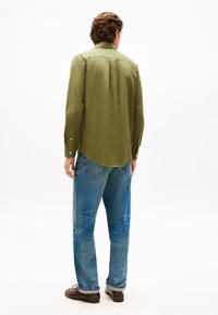 Πουκάμισο μακρυμάνικο σε ελιά πράσινο με στρογγυλεμένη βάση και μανσέτες με κουμπιά, συνδυασμένο με ανοιχτόχρωμο μπλε τζιν και καφέ παπούτσια, βλέποντας από πίσω.
