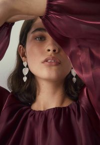 Junge Frau trägt eine durchsichtige burgunderfarbene Bluse und hängende Ohrringe in Form von drei Herzchen aus Kristall, mit einem Arm über ihrem Kopf erhoben.