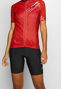 Czerwona koszulka kolarska z krótkim rękawem, ozdobiona białymi wzorami serc i z zamkiem z przodu; zestawiona z czarnymi szortami z wkładkami.